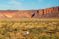A white truck travels along a dirt road through a vast Australian outback landscape, with red rock cliffs and scattered greenery under a bright blue sky.